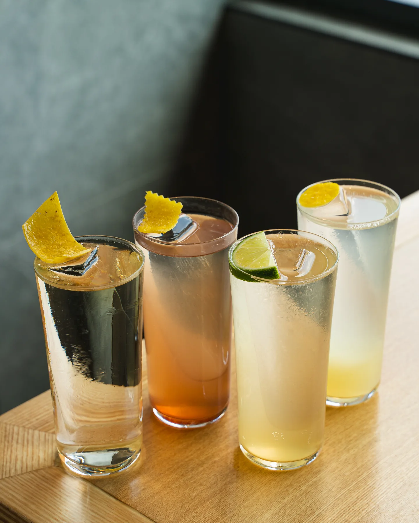 Four tall highballs over cylinders of clear ice, two with twisted lemon peel, on a wood table at Genki Izakaya