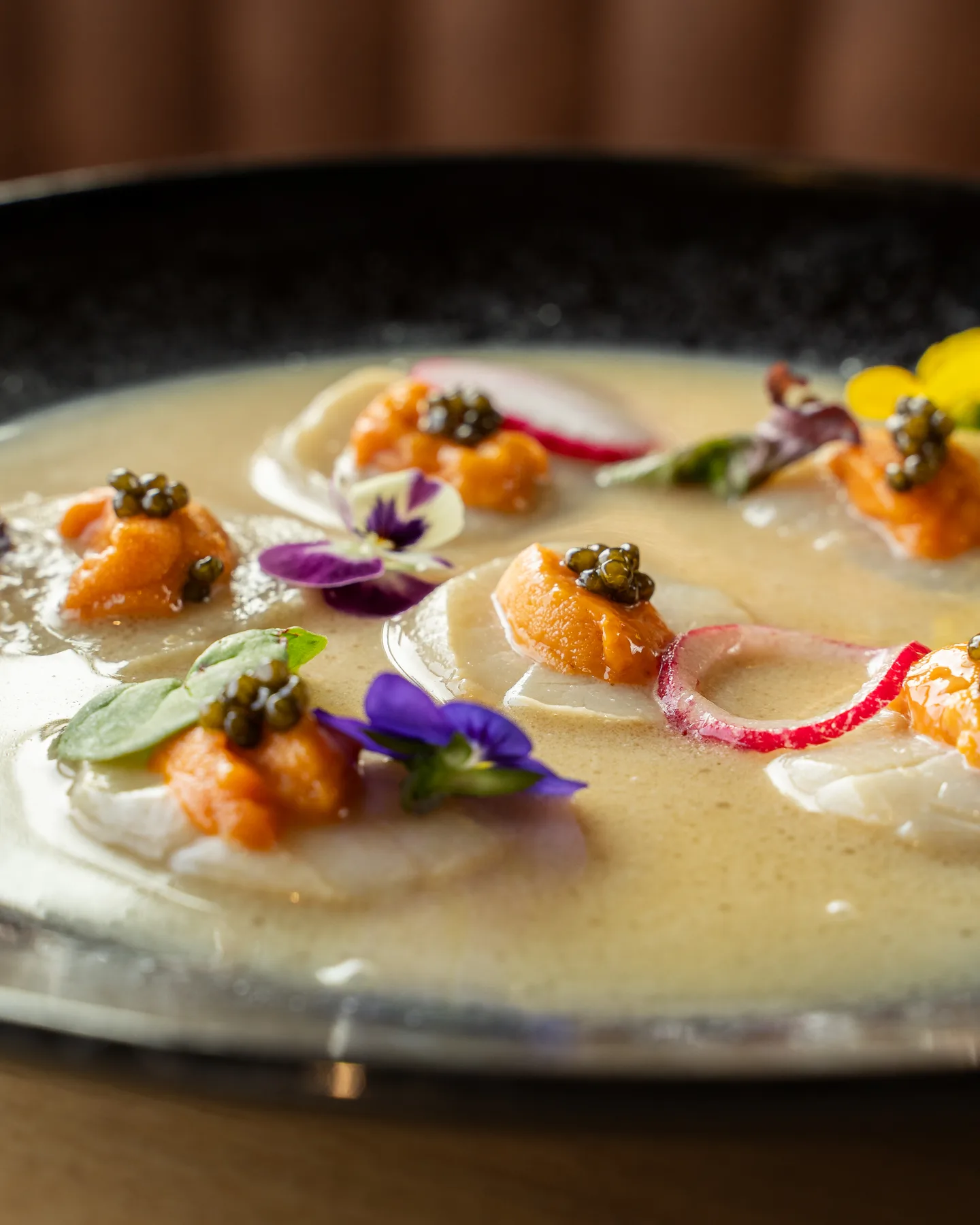 Thin raw scallop slices topped with sea urchin and caviar, with pansies, in a dark speckled bowl at Genki Izakaya