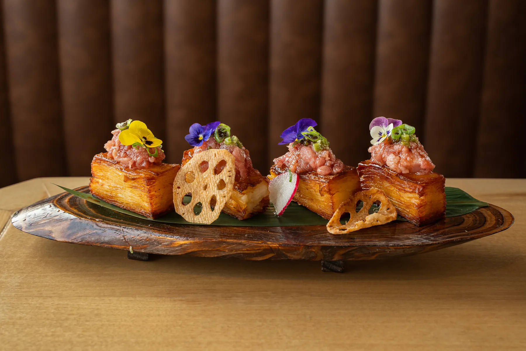 Four golden layered crispy cubes topped with chopped raw tuna, scallion and edible pansies on a dark wood board at Genki Izakaya in Fairfax, VA